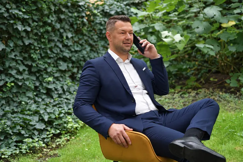 Fehér József öltönyben, sárga széken ül a kertben és telefonál